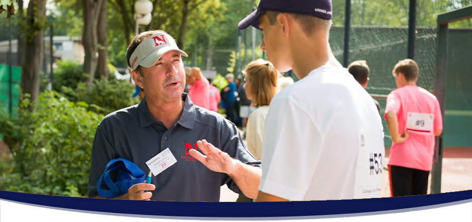 Male college coach in a grey polo shirt talking to a male tennis player in a white shirt