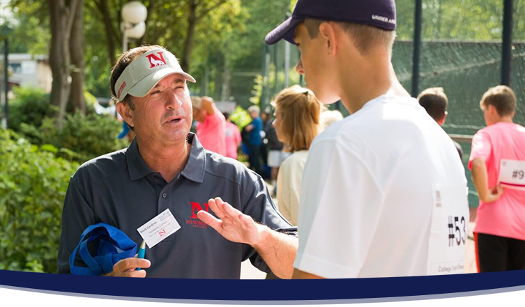 Male college coach in a grey polo shirt talking to a male tennis player in a white shirt